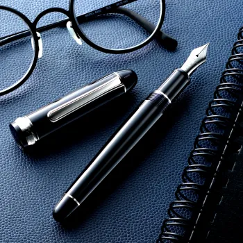 Pluma abierta sobre una mesa junto a cuaderno y gafas redondas